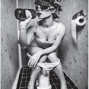 May include: A woman wearing sunglasses and hair rollers sits on a toilet and drinks from a bottle.