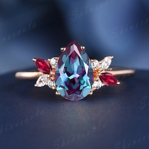 May include: A pear-shaped ring with a large, iridescent blue-green gemstone surrounded by smaller clear gemstones. The ring also features two red gemstones on either side of the main stone.