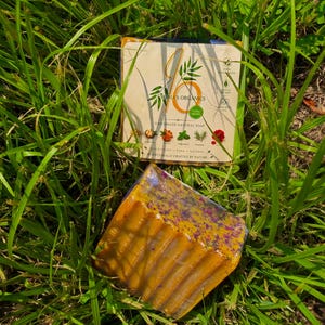 May include: A rectangular bar of handmade natural soap with a golden-brown hue and purple floral inclusions, wrapped in clear plastic. Behind it sits a beige paper label featuring the text Jonia's Organics, botanical illustrations, and icons.