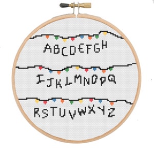 May include: A white embroidery hoop with a cross-stitch design of the alphabet in black letters with a string of colourful lights above each row of letters. The letters are A-H, I-Q, and R-Z.