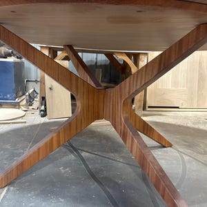 May include: A wooden table featuring a round top and a distinctive starburst base. The base is constructed from angled wooden supports converging at the centre. The table is crafted from light brown wood, likely in a workshop environment.