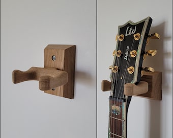 May include: Wooden guitar wall mount. The mount is made of light brown wood and is designed to hold a guitar. The guitar headstock is black with gold tuning pegs. The mount is attached to a white wall.