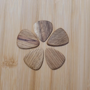 Può includere: Cinque plettri per chitarra in legno disposti a cerchio. I plettri sono realizzati con vari tipi di legno, che mostrano diversi motivi di venature e sfumature di marrone. Lo sfondo è una superficie di legno di colore chiaro.