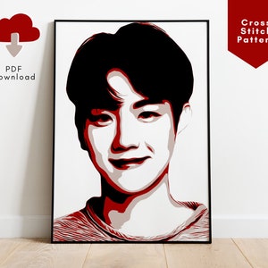 May include: A red and white cross stitch pattern of a person's face. The person has short black hair and is smiling. The pattern is framed in a black frame.