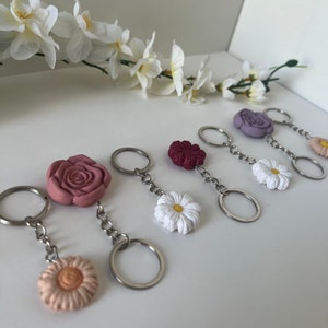 May include: A set of six keychains featuring a variety of flower designs. The keychains are made of metal and feature a daisy and a rose charm. The charms are available in pink, white, purple, and red.