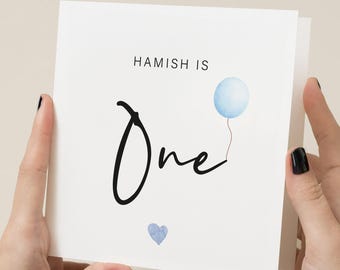 Op de afbeelding: Een witte kaart met de tekst "Hamish is One" in zwarte schrijfletters. Rechts van de tekst staat een blauwe waterverfballon met een touwtje. Een klein blauw hartje staat onderaan de kaart.
