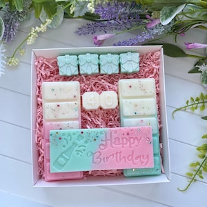 Puede incluir: Una caja de regalo blanca llena de fundidos de cera perfumada rosa y verde. Los fundidos tienen forma de pequeñas cajas de regalo, un número "40" y una barra que dice "Feliz cumpleaños".
