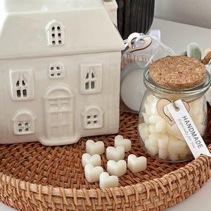 May include: A white ceramic house-shaped candle warmer with window cutouts sits on a woven tray. A glass jar with a cork lid and heart-shaped tag contains heart-shaped wax melts. The tag reads "HANDMADE, Especially for You."