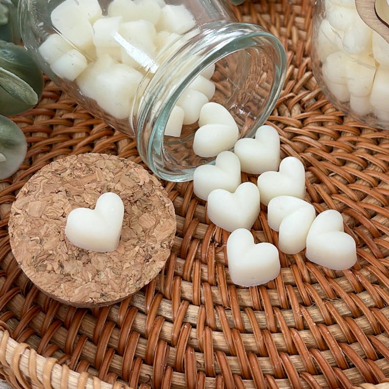 May include: Off-white heart-shaped wax melts are scattered on a wicker tray. A glass jar is tipped over, spilling more melts. A cork coaster holds a single heart-shaped melt. The melts are likely for use in a wax warmer, for fragrance.