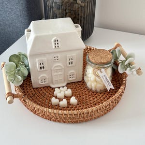 May include: A white ceramic house-shaped wax warmer with cut-out windows and a door sits on a round woven tray. A small glass jar filled with heart-shaped wax melts and a cork lid is next to the warmer. The tray has wooden handles.