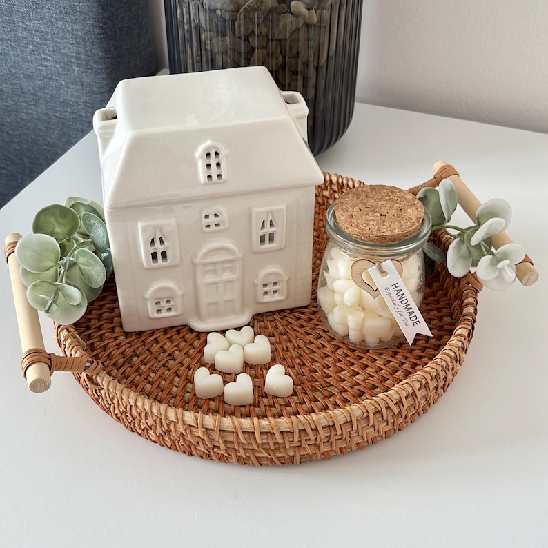 May include: A white ceramic house-shaped wax warmer with cut-out windows and a door sits on a round woven tray. A small glass jar filled with heart-shaped wax melts and a cork lid is next to the warmer. The tray has wooden handles.