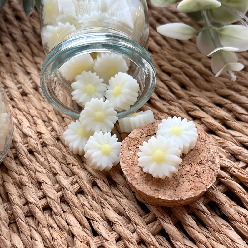 May include: A glass jar, tipped over, contains daisy-shaped wax melts. The white melts have yellow centres. Some are on a cork coaster. The background is a woven, light brown surface.