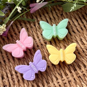 Puede incluir: Cuatro jabones con forma de mariposa en tonos pastel rosa, verde, amarillo y morado. Cada jab&oacute;n tiene un centro brillante y est&aacute; dispuesto sobre una superficie de mimbre tejido. Elementos florales decorativos en el fondo.