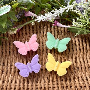 Puede incluir: Cuatro adornos con forma de mariposa en tonos pastel: rosa, verde, lila y amarillo. Cada mariposa tiene un centro brillante y est&aacute; colocada sobre un fondo tejido con elementos florales.