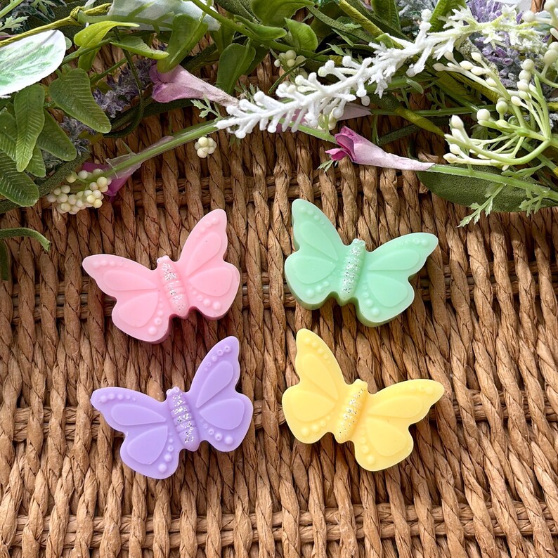 Puede incluir: Cuatro adornos con forma de mariposa en tonos pastel: rosa, verde, lila y amarillo. Cada mariposa tiene un centro brillante y est&aacute; colocada sobre un fondo tejido con elementos florales.