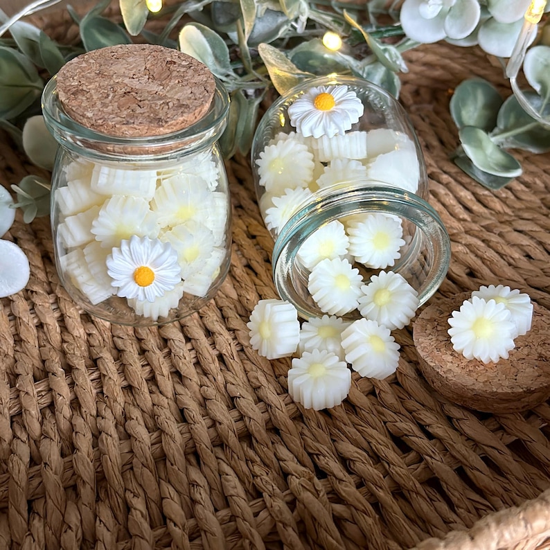 May include: Two small glass jars filled with white daisy-shaped wax melts. One jar has a cork lid, and the other is open, with melts spilling out. Each melt has a yellow centre. The jars sit on a woven brown surface.