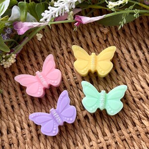Puede incluir: Cuatro figuras de cera con forma de mariposa en rosa, amarillo, lila y verde. Cada mariposa tiene detalles de purpurina. Las figuras est&aacute;n dispuestas sobre una superficie tejida marr&oacute;n, con elementos florales.