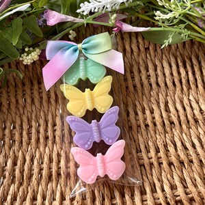 Puede incluir: Una bolsa transparente de celof&aacute;n contiene cuatro jabones con forma de mariposa en colores pastel: verde, amarillo, morado y rosa. Un lazo de cinta arco&iacute;ris pastel est&aacute; atado en la parte superior. Los jabones est&aacute;n sobre un fondo de mimbre tejido con acentos florales.