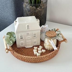 May include: A woven rattan tray with wooden handles displays a white ceramic house-shaped wax warmer, a glass jar of heart-shaped wax melts, and artificial greenery. The tray sits on a white surface.