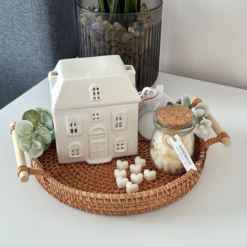 May include: A woven rattan tray with wooden handles displays a white ceramic house-shaped wax warmer, a glass jar of heart-shaped wax melts, and artificial greenery. The tray sits on a white surface.