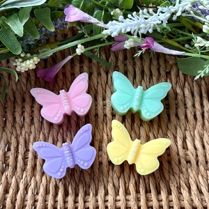 Puede incluir: Cuatro objetos con forma de mariposa en tonos pastel: rosa, verde menta, lila y amarillo. Cada mariposa tiene un centro brillante. Est&aacute;n dispuestas sobre un fondo tejido, con decoraciones florales.
