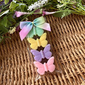 Puede incluir: Una bolsa transparente de celof&aacute;n contiene cuatro jabones con forma de mariposa en colores pastel: verde, amarillo, lila y rosa. Los jabones est&aacute;n decorados con peque&ntilde;os puntos y purpurina. Una cinta de lazo multicolor est&aacute; sujeta a la parte superior de la bolsa. El fondo es una cesta tejida.