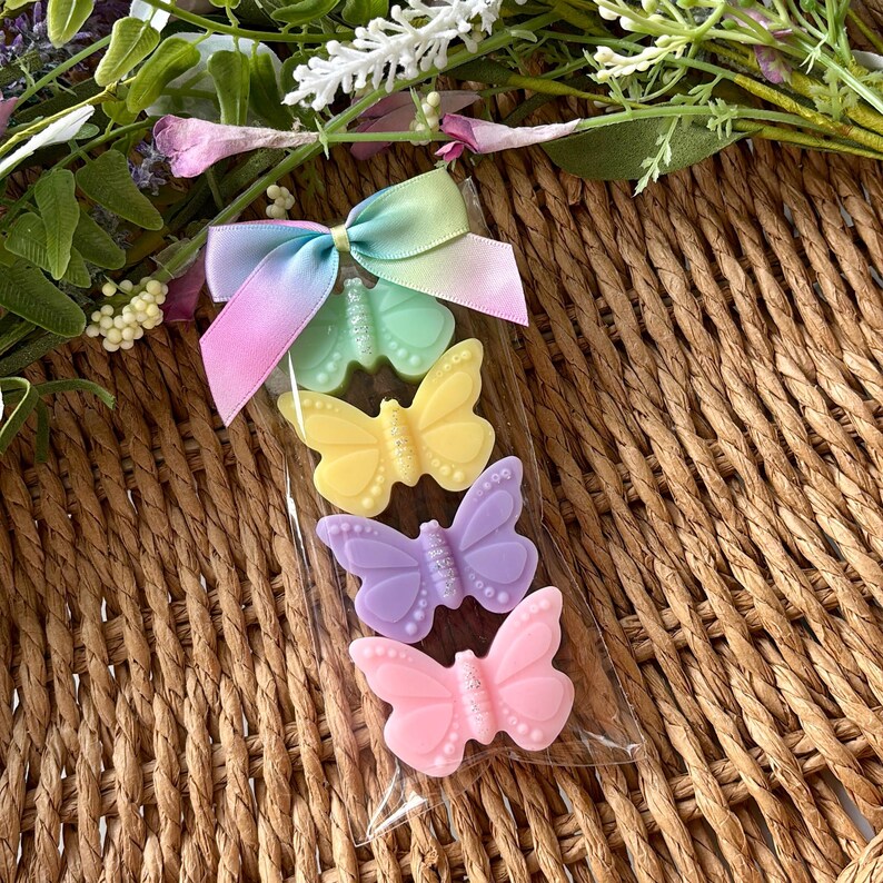 Puede incluir: Una bolsa transparente de celof&aacute;n contiene cuatro jabones con forma de mariposa en colores pastel: verde, amarillo, lila y rosa. Los jabones est&aacute;n decorados con peque&ntilde;os puntos y purpurina. Una cinta de lazo multicolor est&aacute; sujeta a la parte superior de la bolsa. El fondo es una cesta tejida.