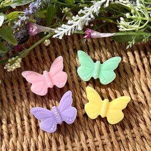 Puede incluir: Cuatro decoraciones con forma de mariposa en colores pastel: rosa, verde, morado y amarillo, cada una con un centro brillante. Las mariposas est&aacute;n dispuestas sobre una superficie marr&oacute;n tejida, con acentos florales en el fondo.