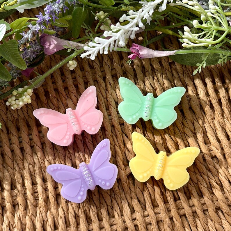 Puede incluir: Cuatro decoraciones con forma de mariposa en colores pastel: rosa, verde, morado y amarillo, cada una con un centro brillante. Las mariposas est&aacute;n dispuestas sobre una superficie marr&oacute;n tejida, con acentos florales en el fondo.