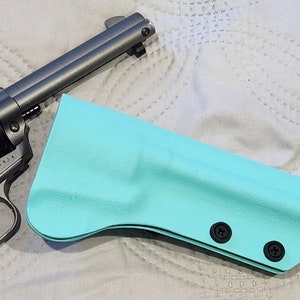 May include: A silver revolver and a turquoise holster are displayed on a white, quilted surface. The revolver has a black grip and a cylinder. The holster is made of a smooth material and has two black screws.