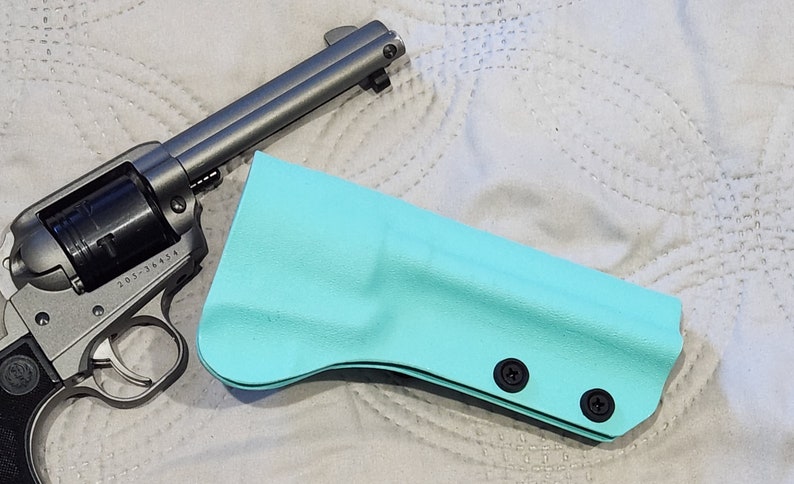 May include: A silver revolver and a turquoise holster are displayed on a white, quilted surface. The revolver has a black grip and a cylinder. The holster is made of a smooth material and has two black screws.