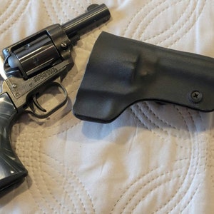 Può includere: Un revolver nero con grilletto argentato e impugnatura grigia marmorizzata, accanto a una fondina nera. Il revolver ha un tamburo e una canna. L'impugnatura presenta un motivo a spirale grigio e bianco. La fondina è modellata sulla forma della pistola.