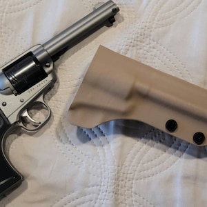 May include: A silver revolver with a black grip and a tan holster. The revolver has a cylinder and a trigger. The holster is made of a rigid material and has three black rivets.