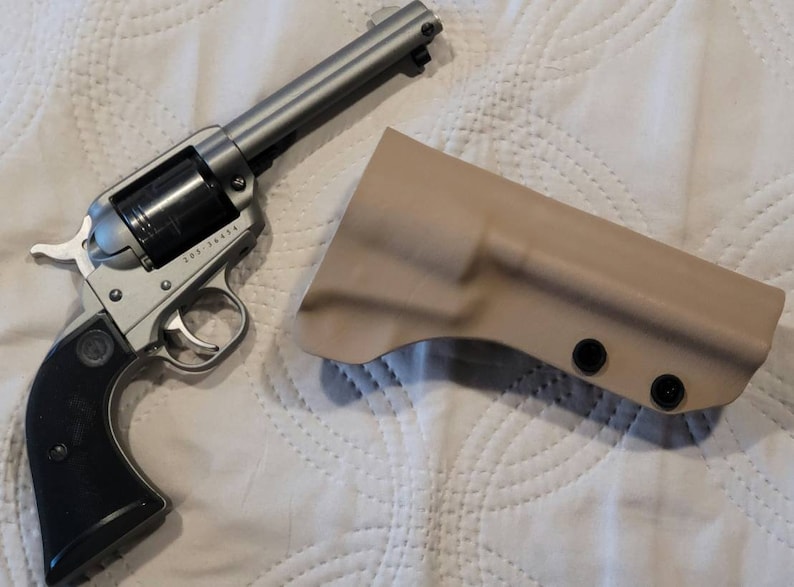 May include: A silver revolver with a black grip and a tan holster. The revolver has a cylinder and a trigger. The holster is made of a rigid material and has three black rivets.