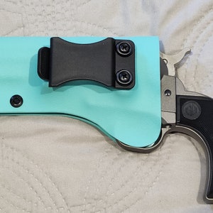 May include: A turquoise pistol holster with a black belt clip and black hardware. A silver and black handgun is partially visible, with a black grip and silver trigger and hammer.