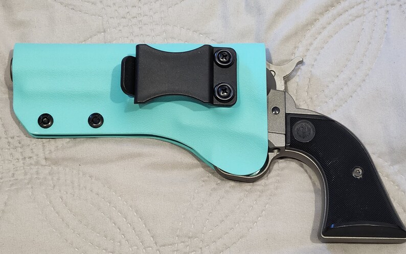 May include: A turquoise pistol holster with a black belt clip and black hardware. A silver and black handgun is partially visible, with a black grip and silver trigger and hammer.