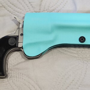May include: A single-action revolver with a black grip and a silver frame is partially encased in a light blue holster. The holster is secured with two black screws. The Ruger logo is visible on the grip.