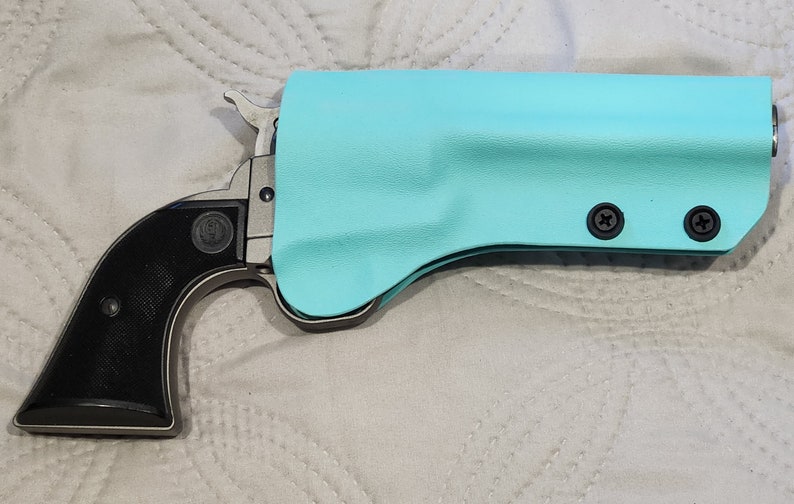May include: A single-action revolver with a black grip and a silver frame is partially encased in a light blue holster. The holster is secured with two black screws. The Ruger logo is visible on the grip.