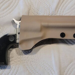 May include: A black and silver handgun with a black grip is partially inserted into a tan holster. The holster is secured with black screws. The handgun is a single-action revolver.
