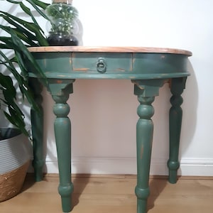 May include: A small, green painted wooden table with a single drawer and turned legs. The tabletop is natural wood. A glass terrarium with plants sits on top. The table is approximately 76 cm wide.