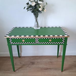 May include: A green wooden desk with two drawers featuring a black and white chequered border along the top edge and drawer fronts. The desk has four tapered legs and two small gold-toned circular knobs. A clear glass vase with white flowers sits on top.