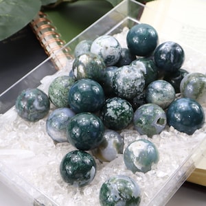 May include: A collection of green and white moss agate spheres in a clear tray. The spheres are arranged in a random pattern and are surrounded by white crystals.