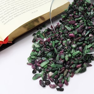 May include: A close-up of a glass bowl filled with green and pink gemstones. The bowl is tipped over, spilling the gemstones onto a white surface.