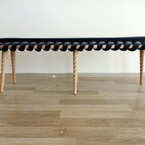 Può includere: Una panca imbottita nera con un design intrecciato e quattro gambe in legno. Le gambe sono intrecciate e hanno una finitura in legno naturale.