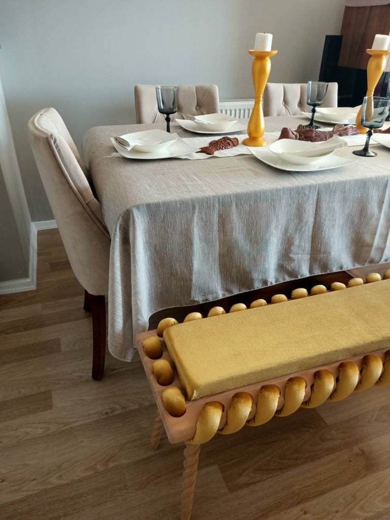 K&ouml;nnte beinhalten: Ein Esszimmer mit einem gedeckten Tisch. Der Tisch ist mit einer hellgrauen Tischdecke bedeckt und mit wei&szlig;en Tellern, Gl&auml;sern und dekorativen Gegenst&auml;nden gedeckt. Vor dem Tisch befindet sich eine gelbe Bank mit gelbem Kissen.