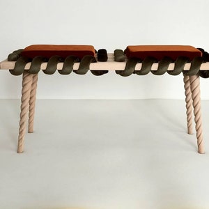 Può includere: Una panca in legno con un design unico. La seduta è realizzata con un materiale intrecciato simile a una corda e ha due cuscini. Le gambe sono intrecciate e hanno una finitura rosa chiaro.