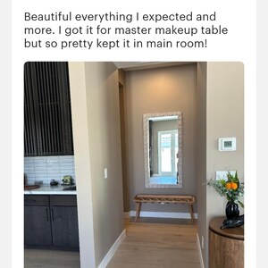 May include: A hallway with light brown walls and wood floors. A rectangular mirror with a light brown frame hangs on the wall. A wooden bench with a woven seat sits in front of the mirror. A small vase with orange flowers sits on a round wooden table.