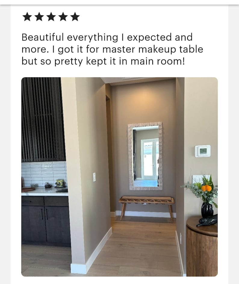 May include: A hallway with light brown walls and wood floors. A rectangular mirror with a light brown frame hangs on the wall. A wooden bench with a woven seat sits in front of the mirror. A small vase with orange flowers sits on a round wooden table.