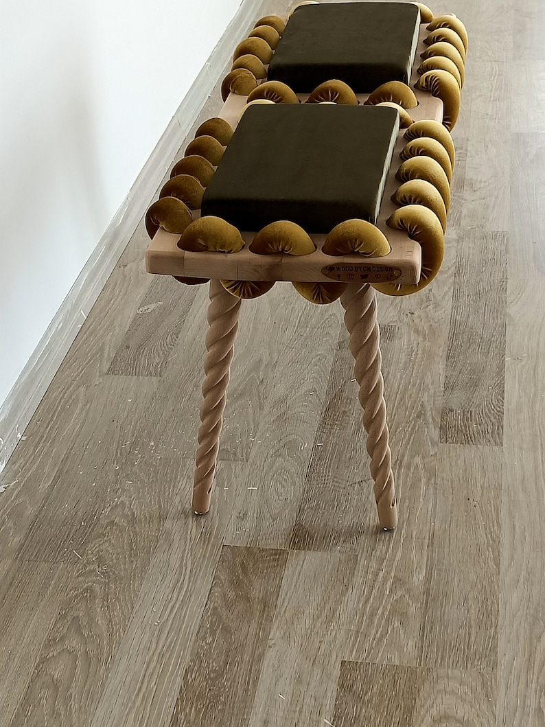 May include: A wooden bench with two dark green cushions and a gold velvet rope design. The bench has four twisted legs and a label that reads "WOOD BY RYK DESIGN".