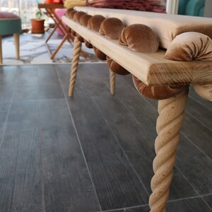 May include: A wooden bench with a beige cushion and rope legs. The cushion is decorated with brown velvet buttons.